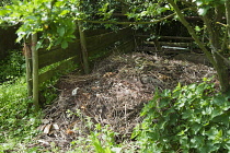 A compost heap made with wooden planks and posts in a wild area with nettles growing nearby.