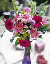 Lily, Lilium 'Star Gazer' with globe thistles, green chrysanthemums, pink gerberas and roses. arranged in a vase on a white table outdoors.