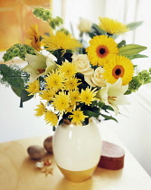 Chrysanthemum, yellow Gerberas, cream Roses and Easter lily, Lilium longifolium, with greenery in a vase on a small wooden table.