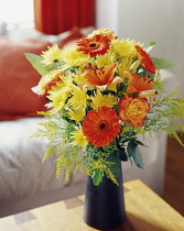 Daisy, Chrysanthemum, Floral arrangment with orange Gerberas, Roses and Oriental Lilies with greenery in a black vase on a small wooden table.