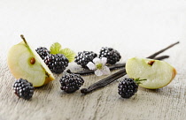 Blackberry, Rubus cultivar. Several berries arranged with flowers and leaves and pieces of apple, plus vanilla pods on pale, distressed, wooden background. Selective focus.