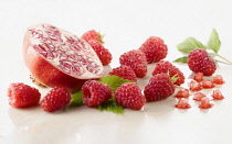 Raspberry, Rubus idaeus cultivar and Pomegranate, Punica cultivar cut in half surrounded with several raspberries and leaves on white marble. Selective focus.