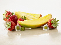 Mango, Mangifera indica. Two slices arranged with strawberries, Fragaria cultivar and flowers, on white marble. Selective focus.